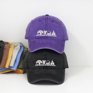 May include: Two baseball caps, one purple and one black, stacked on top of each other. Each cap features a white graphic of a tree, Mickey Mouse, a golf ball, and a castle. The caps are made of a soft, worn material.
