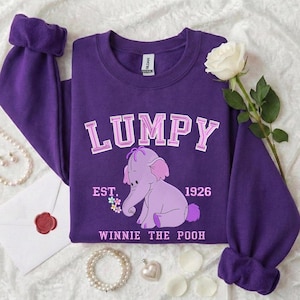 May include: A purple sweatshirt featuring the word "LUMPY" in white and pink lettering above a cartoon elephant. The text "EST. 1926" and "WINNIE THE POOH" are also printed on the shirt. A white rose and jewelry are in the background.