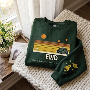 May include: A dark green sweatshirt with a retro space-themed design. The design includes a sunset, planets, and the word "ERID". The sleeve features a rocket and planet graphic. The text "First Planet from Eridani 40" is also visible.
