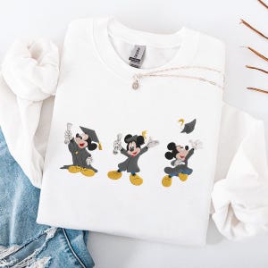 May include: White sweatshirt featuring embroidered Mickey Mouse characters in graduation attire. The design shows Mickey in a graduation gown, holding a diploma, and throwing his cap. A silver necklace with a pendant is also visible.
