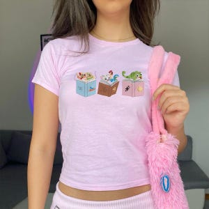 May include: A light pink t-shirt featuring three open books with cartoon animals on them. The books are blue, brown, and pink. A pink fluffy bag is held in the image.