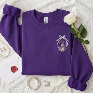 May include: A purple sweatshirt with a crew neck and long sleeves. The cuffs are folded over. A small embroidered design of a castle is on the front. A white rose and pearl jewelry are next to the sweatshirt.