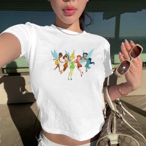 May include: White crop top featuring a colorful graphic of six fairies with wings. The fairies are depicted in various poses and outfits, with colors including yellow, orange, green, red, and blue. The shirt has a round neckline and short sleeves.