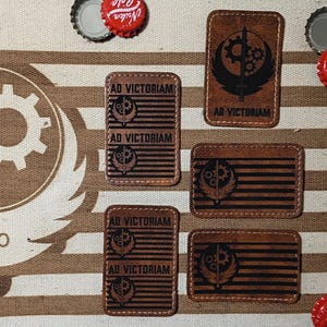Peut inclure: Plusieurs écussons en cuir marron avec le texte "AD VICTORIAM" et un emblème noir avec un engrenage et des ailes. Les écussons sont rectangulaires et ont une bordure cousue. Des capsules de bouteilles sont éparpillées.