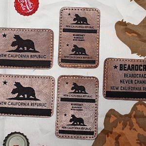 Peut inclure: Plusieurs écussons en cuir marron avec l'inscription "New California Republic" et une silhouette d'ours. Certains écussons affichent également "Bearocracy Never Changes". Les écussons sont rectangulaires avec des bords cousus.