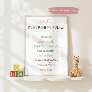 May include: A framed print with the text "LUCA'S PLAYROOM RULES" in colorful letters. The rules include "Be Kind," "Laugh Loudly," and "Have Fun." A small giraffe plush toy and ABC blocks sit on a white shelf below the print.