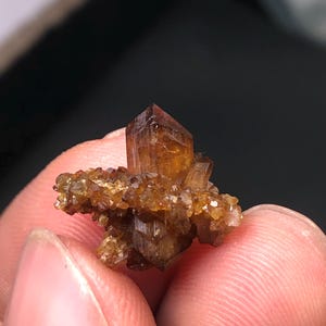May include: A cluster of brown crystals with a prominent, sharply defined central crystal. The crystals have a warm, amber hue and are set against a blurred background. The mineral specimen is held between fingers.