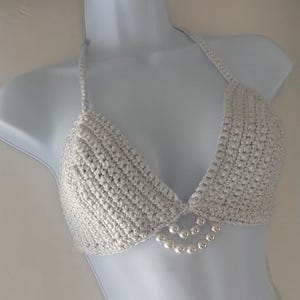 Könnte beinhalten: Silberfarbenes, gehäkeltes Bikini-Oberteil mit Neckholder-Design. Die Dreiecks-Cups sind durch eine kleine, perlenbesetzte Verzierung verbunden. Der untere Teil des Oberteils hat drei Stränge kleiner, runder Perlen.