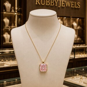 Op de afbeelding: Een gouden ketting met een grote, rechthoekige, gefacetteerde roze edelsteen hanger in een gouden zetting. De ketting wordt gepresenteerd op een neutraal gekleurde sieradenbuste, met de merknaam RUBBYJEWELS zichtbaar op de achtergrond.