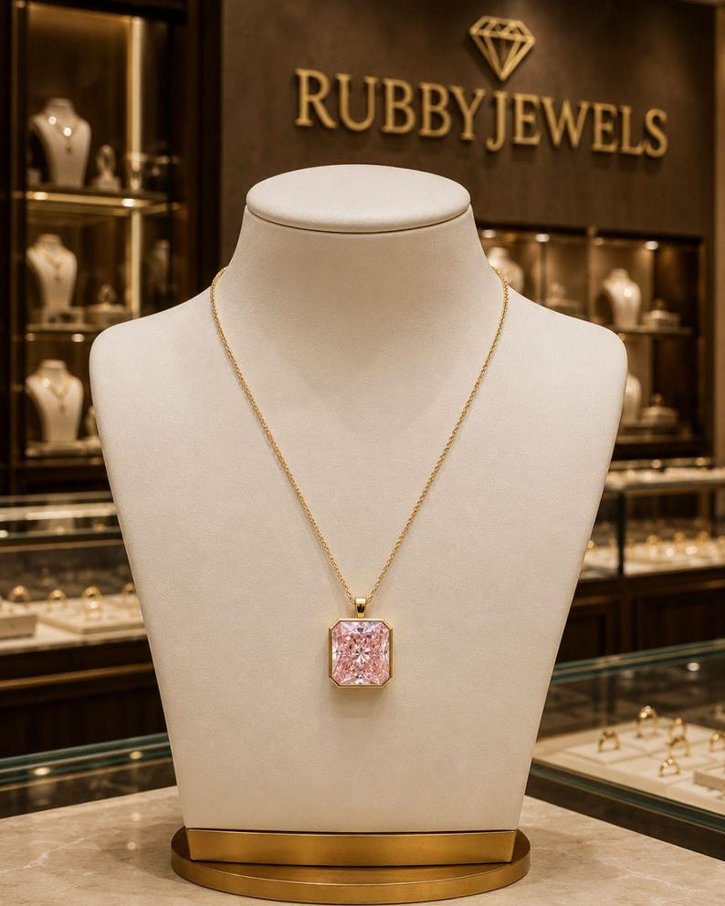 Op de afbeelding: Een gouden ketting met een grote, rechthoekige, gefacetteerde roze edelsteen hanger in een gouden zetting. De ketting wordt gepresenteerd op een neutraal gekleurde sieradenbuste, met de merknaam RUBBYJEWELS zichtbaar op de achtergrond.