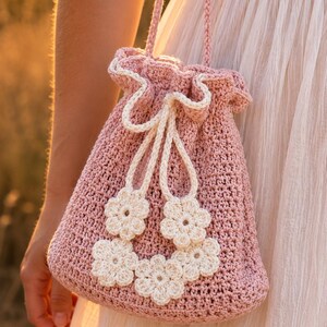 Peut inclure: Un sac rose en crochet avec des fleurs blanches. Le sac a un haut à volants et une fermeture à cordon blanche. Le sac est tenu par une sangle et comporte des fleurs blanches en crochet.