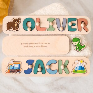 Op de afbeelding: Twee houten naam puzzels met de namen "OLIVER" en "JACK" in kleurrijke letters. Elke puzzel heeft decoratieve elementen, waaronder een beer, een dinosaurus, een trein en een berglandschap. De middelste puzzel heeft de tekst: "For our sweetest little one - with love, Auntie Emmy."
