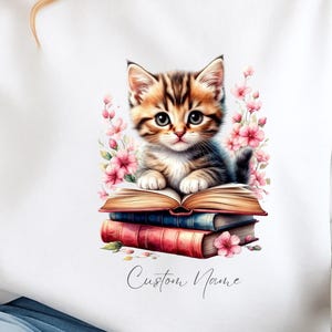 May include: White sweatshirt with a watercolour illustration of a kitten on a stack of books, framed by pink flowers. The cursive text "Custom Name" is below the books. The design is centrally placed on the front of the garment.