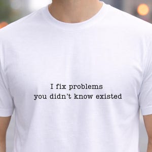 Könnte beinhalten: Weißes T-Shirt mit dem Text "I fix problems you didn't know existed" in schwarzer Schrift. Das Shirt hat kurze Ärmel und einen Rundhalsausschnitt. Der Hintergrund ist unscharf, was auf eine Außenumgebung hindeutet.
