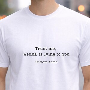 May include: White t-shirt with the text "Trust me, WebMD is lying to you" and "Custom Name" printed in black. The shirt is made of a soft material and is being worn.