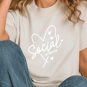 Puede incluir: Una camiseta beige con las palabras "Social Worker" en escritura blanca, con un corazón y pequeños corazones. La camiseta tiene mangas cortas y se combina con unos vaqueros azules.
