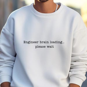 Puede incluir: Una sudadera blanca con el texto "Engineer brain loading... please wait" en negro. La sudadera tiene cuello redondo y mangas largas. La persona lleva un reloj.