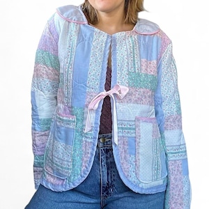 May include: A quilted jacket in pastel colors, featuring a patchwork design with floral and solid patterns. The jacket has a rounded collar and pockets, and is tied with a pink ribbon. The jacket is a light blue, pink, and green color.
