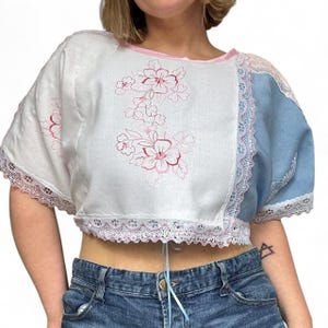 May include: A cropped top with a patchwork design. The top features white fabric with pink floral embroidery, a light blue panel, and pink lace trim. The neckline and hem are trimmed with pink. The top has short sleeves.