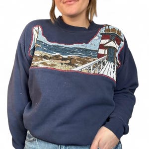 Può includere: Felpa blu navy con un design ricamato dettagliato di un faro su una costa rocciosa. Il faro presenta strisce rosse e bianche, con un oceano blu e rocce beige. La felpa ha un girocollo e maniche lunghe.
