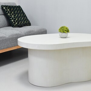 May include: A white, kidney-shaped coffee table with a rounded base. A small potted plant sits on top. A gray sofa with a gold and black patterned pillow is in the background.