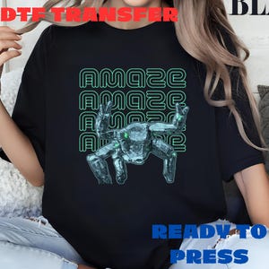 May include: Black t-shirt with a teal graphic of a robot-like figure and the word "AMAZE" repeated three times. The words "READY TO PRESS" are printed in blue at the bottom. The words "DTF TRANSFER" are printed in red at the top.
