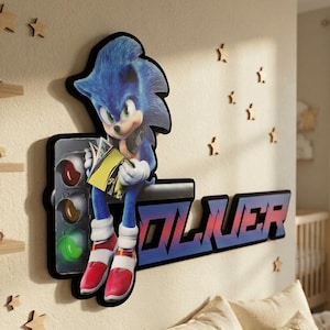 May include: A wall-mounted sign featuring a blue hedgehog character sitting on a black horizontal bar next to a traffic light. The character holds a yellow book. The name OLIVER is displayed in large, bold, blue and red block lettering.