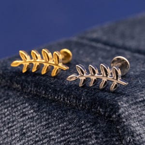 May include: Two leaf-shaped stud earrings. One is gold-toned, the other is silver-toned. Both earrings have a post backing. The earrings are displayed on a dark blue textured surface.