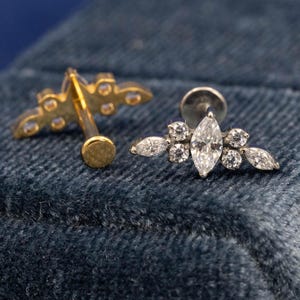 May include: Two stud earrings with a floral design. One earring is gold-toned with small clear stones. The other earring is silver-toned with a marquise-cut center stone and surrounding clear stones. The earrings are displayed on a blue textured surface.