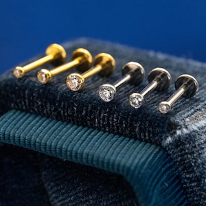 May include: A selection of six small stud earrings. Three are gold-coloured with clear, round gemstones, and three are silver-coloured with similar gemstones. The earrings are displayed on a textured, dark blue surface, with a solid blue background.