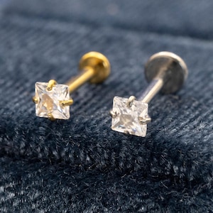 May include: A pair of small stud earrings, each featuring a square, clear gemstone. One earring has a gold post and backing, the other silver. They are set against a dark blue, textured background.
