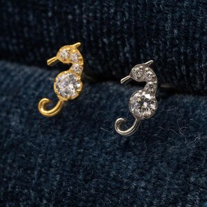 May include: Two seahorse-shaped earrings. One is gold-toned, the other silver-toned. Both earrings feature clear, sparkling gemstones. The seahorses are set against a dark blue, textured background.