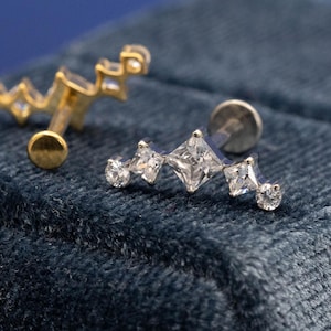 May include: Close-up of two stud earrings. One is gold-toned with a curved design and small clear stones. The other is silver-toned with a row of clear, square and round-cut stones. Both are on a dark blue velvet surface.