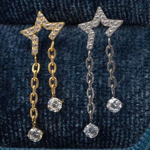 May include: Two star-shaped earrings, one gold and one silver, each with a chain and a dangling clear, round stone. The stars are set with small, clear stones. The earrings are shown against a dark blue backdrop.