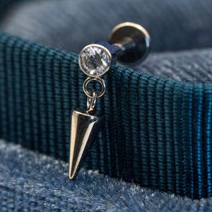 May include: A silver labret stud featuring a clear, round gemstone and a dangling, polished, silver cone. The jewellery is set against a dark blue backdrop, accentuating the sparkle of the gem and the sleek design.