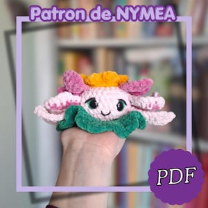 May include: A handmade crochet flower in shades of pink, yellow, and green. The flower has a smiling face and is held in a hand. The image includes the text "Patron de NYMEA" and "PDF".