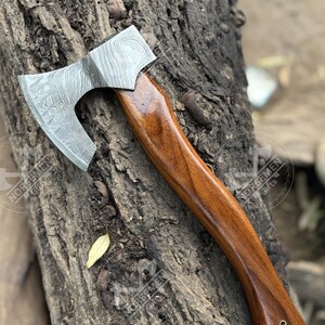 May include: A Damascus steel axe with a polished wooden handle. The axe head features a distinctive pattern, and the handle is a rich, dark brown. The axe is displayed on a textured, brown surface.