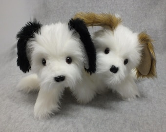 May include: Two plush toy dogs. One dog has black ears, the other has brown ears. Both have white fur, black noses, and dark eyes. The dogs are positioned close together, appearing to be cuddling. The background is a light gray.