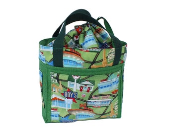 May include: Green fabric lunch bag with a Route 66 diner print. The bag has a drawstring closure and a green handle. The exterior features a pocket and is trimmed with green fabric. The bag is designed to carry food and drinks.