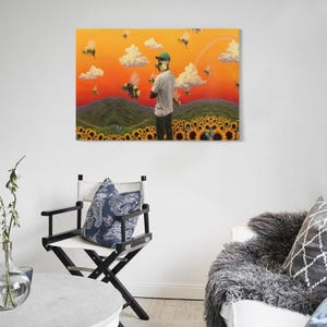 May include: A framed canvas art print depicts a person in a field of sunflowers, surrounded by bees, under a vibrant orange sky with fluffy white clouds. The person is wearing a green cap and a white shirt. The artwork is displayed on a white wall.