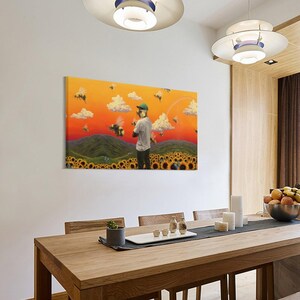 May include: A rectangular canvas art print depicts a person in a field of sunflowers with bees and clouds against an orange sky. The person is wearing a green hat and a white shirt. The artwork is displayed on a white wall in a dining room.
