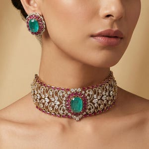 Pode incluir: Uma mulher usando um colar gargantilha dourado e brincos combinando. As joias apresentam pedras centrais ovais de cor esmeralda, rodeadas por pedras vermelhas e transparentes. O colar e os brincos têm um design floral ornamentado.