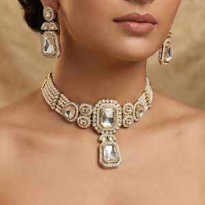 Op de afbeelding: Een goudkleurige sieradenset met een ketting, oorbellen en een hoofdband. De ketting heeft een grote, rechthoekige, heldere stenen hanger. De oorbellen en hoofdband hebben ook vergelijkbare rechthoekige stenen. De sieraden staan tegen een neutrale achtergrond.