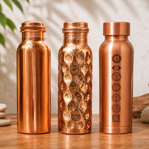 Puede incluir: Tres botellas de agua de cobre con tapas de rosca. Las botellas tienen diferentes diseños: una es lisa, otra tiene un patrón texturizado y la tercera presenta símbolos de chakra grabados. Las botellas se exhiben sobre una superficie de madera.