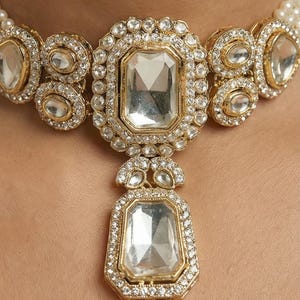 Op de afbeelding: Close-up van een goudkleurige ketting met grote, heldere rechthoekige en ovale edelstenen. De edelstenen zijn omgeven door kleinere, heldere stenen. De ketting is geplaatst tegen een lichte achtergrond en lijkt een statement-stuk te zijn.