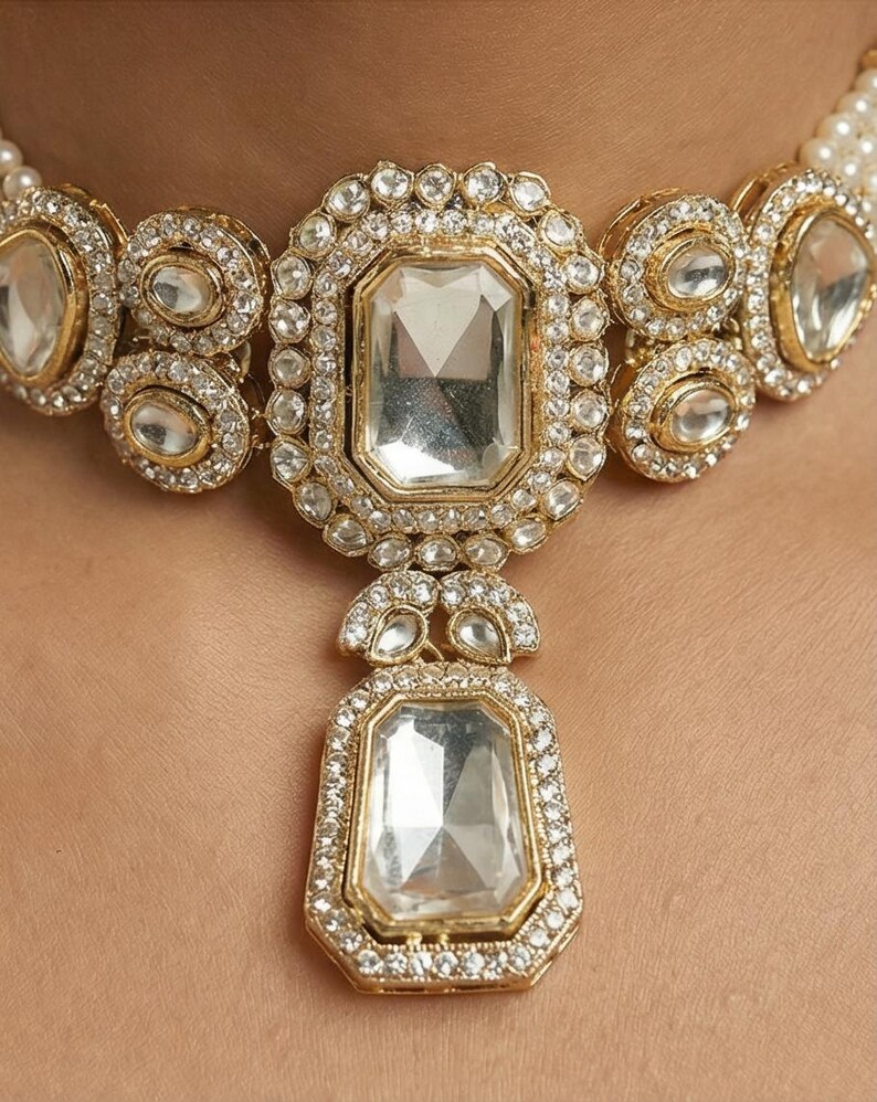 Op de afbeelding: Close-up van een goudkleurige ketting met grote, heldere rechthoekige en ovale edelstenen. De edelstenen zijn omgeven door kleinere, heldere stenen. De ketting is geplaatst tegen een lichte achtergrond en lijkt een statement-stuk te zijn.