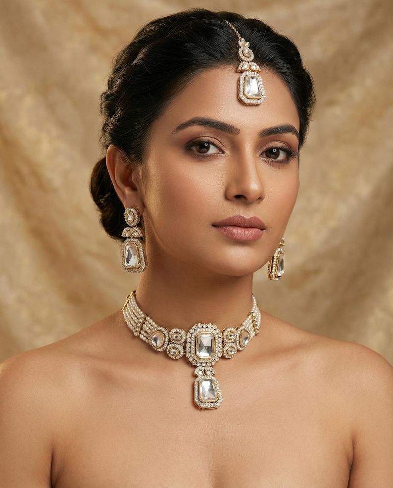 Op de afbeelding: Een goudkleurige sieradenset met een ketting, oorbellen en een hoofdband. De ketting heeft een grote, rechthoekige, heldere stenen hanger. De oorbellen en hoofdband hebben ook vergelijkbare rechthoekige stenen. De sieraden staan tegen een neutrale achtergrond.