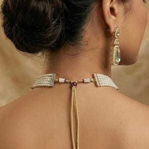 Op de afbeelding: Een close-up van een goudkleurige ketting en bijpassende oorbellen. De ketting heeft een parel- en strassontwerp met een gouden koord. De oorbellen zijn rechthoekig met een heldere steen en gouden accenten. De sieraden worden door een persoon gedragen.