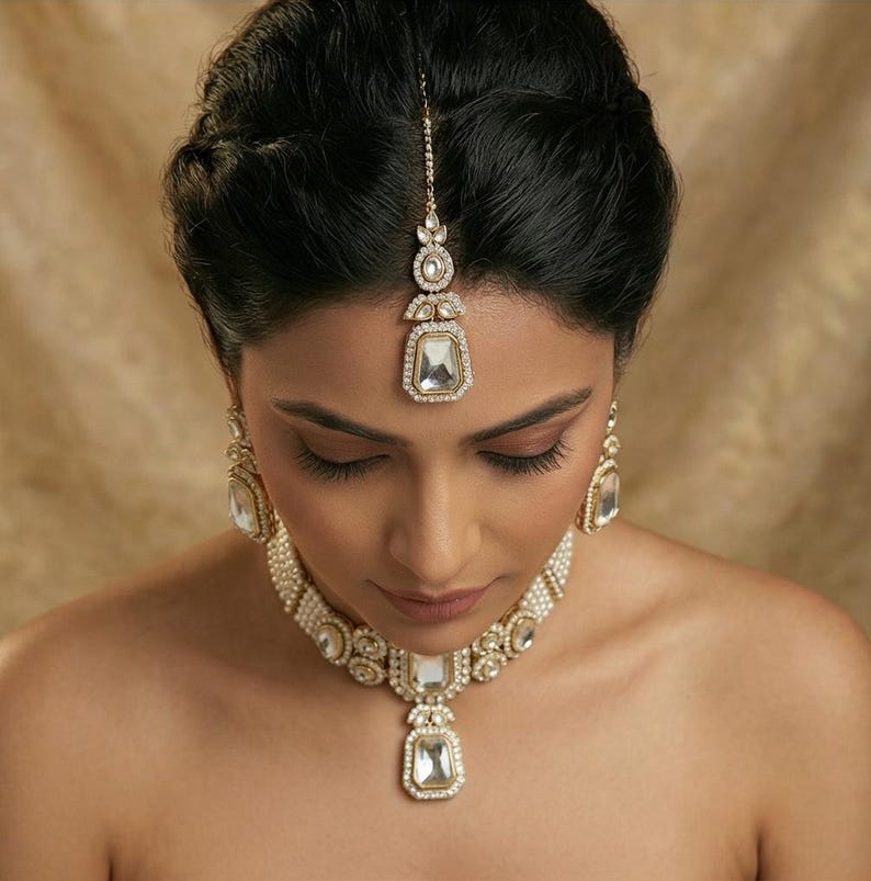 Op de afbeelding: Een vrouw draagt een goudkleurige sieradenset. De set bestaat uit een ketting, oorbellen en een hoofdsieraad, allemaal met grote, heldere, vierkant geslepen stenen en kleinere, ronde stenen. De sieraden zijn geplaatst tegen een neutrale achtergrond.