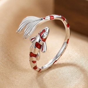 Könnte beinhalten: Ein silberner Ring mit einem Koi-Karpfen-Design. Der Fisch hat rot-weiße Markierungen und einen kleinen Edelstein. Der Ring umschließt den Finger, wobei sich die Schwanzflosse des Fisches nach oben erstreckt. Ein einzigartiges Schmuckstück.